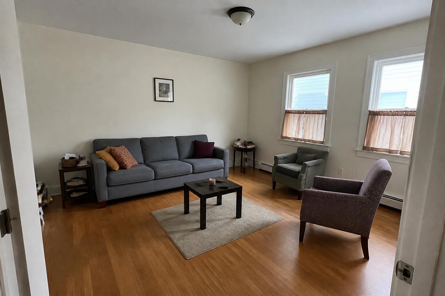 A living room before its Edit: a gray sofa, two mismatched accent chairs, an undersized rug floating in the middle of the floor, short cafe curtains and one small framed print